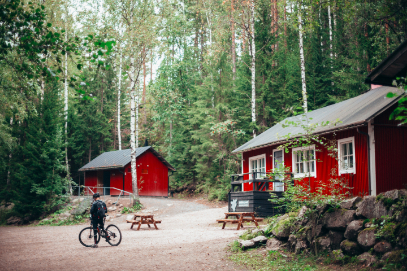 Zwei rote Holzhäuser und ein Fahrradfahrer mittem im Wald.